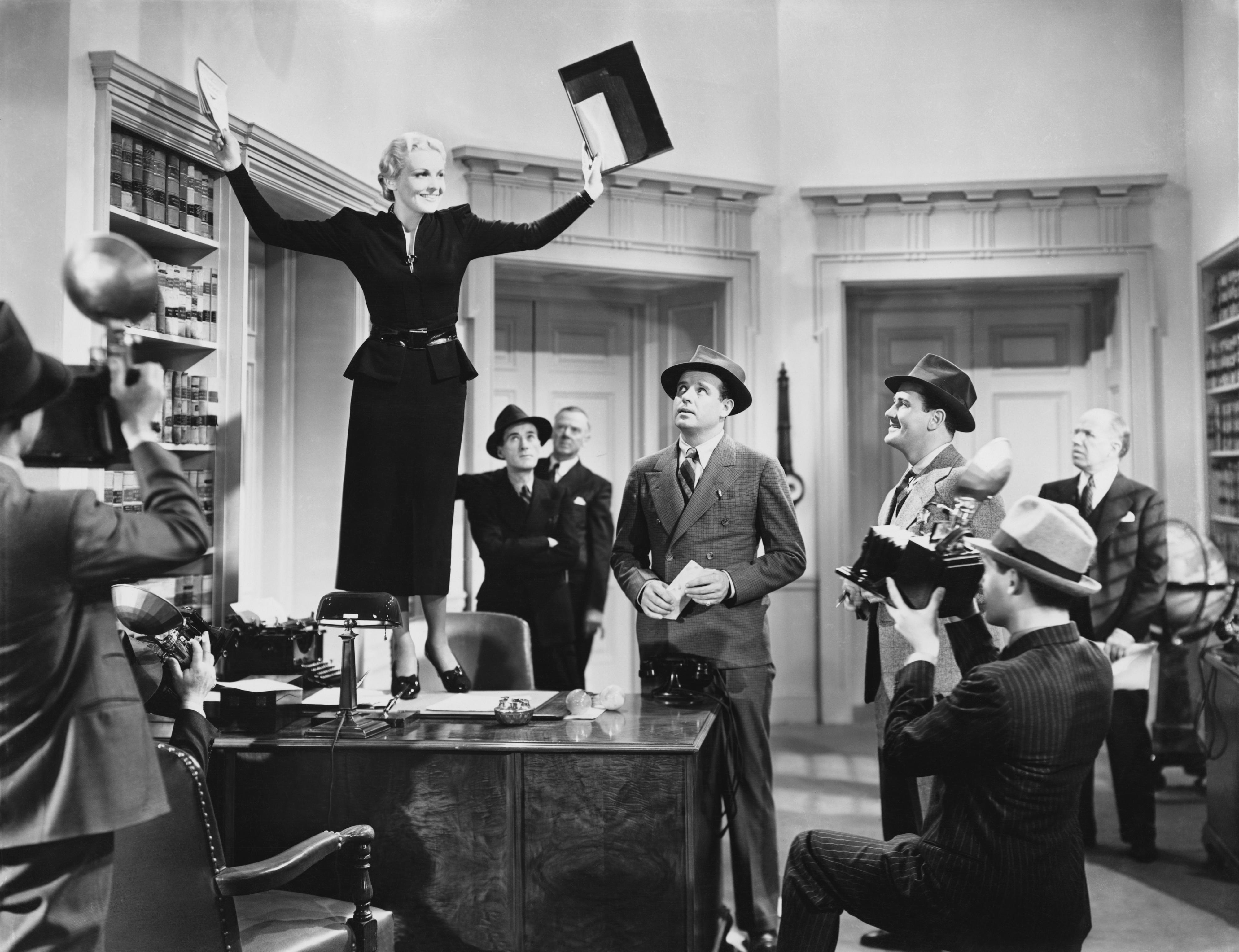 Woman standing on table with hands up surrounded by a group of men in suits