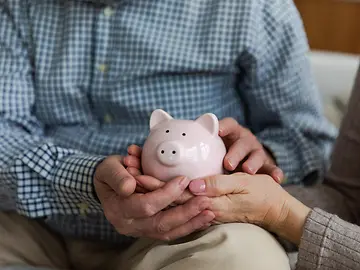 A pink piggy bank, being held by male hands intertwined with female hands that are painted with pink nail varnish.