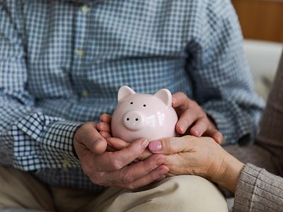 A pink piggy bank, being held by male hands intertwined with female hands that are painted with pink nail varnish.