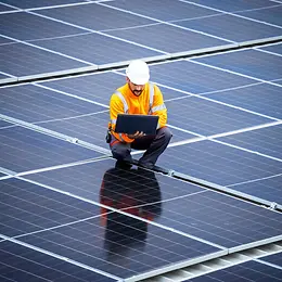 A man in a yellow jacket, blue trousers and a white helmet holds a laptop while crouching in the middle of solar modules.