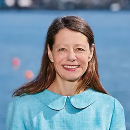 A middle-aged woman with long brown hair and a light blue suit is smiling into the camera