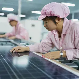 Workers in pink coats and caps inspect solar panels in a brightly-lit factory.