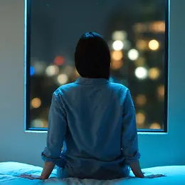Woman sits on bed at night and looks out of the window
