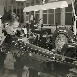 A black and white photo shows a worker operating a mechanical press that is minting a coin.