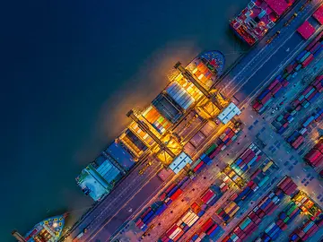 A dock covered in shipping containers in a variety of colours. A ship is docked and is being loaded at dusk.