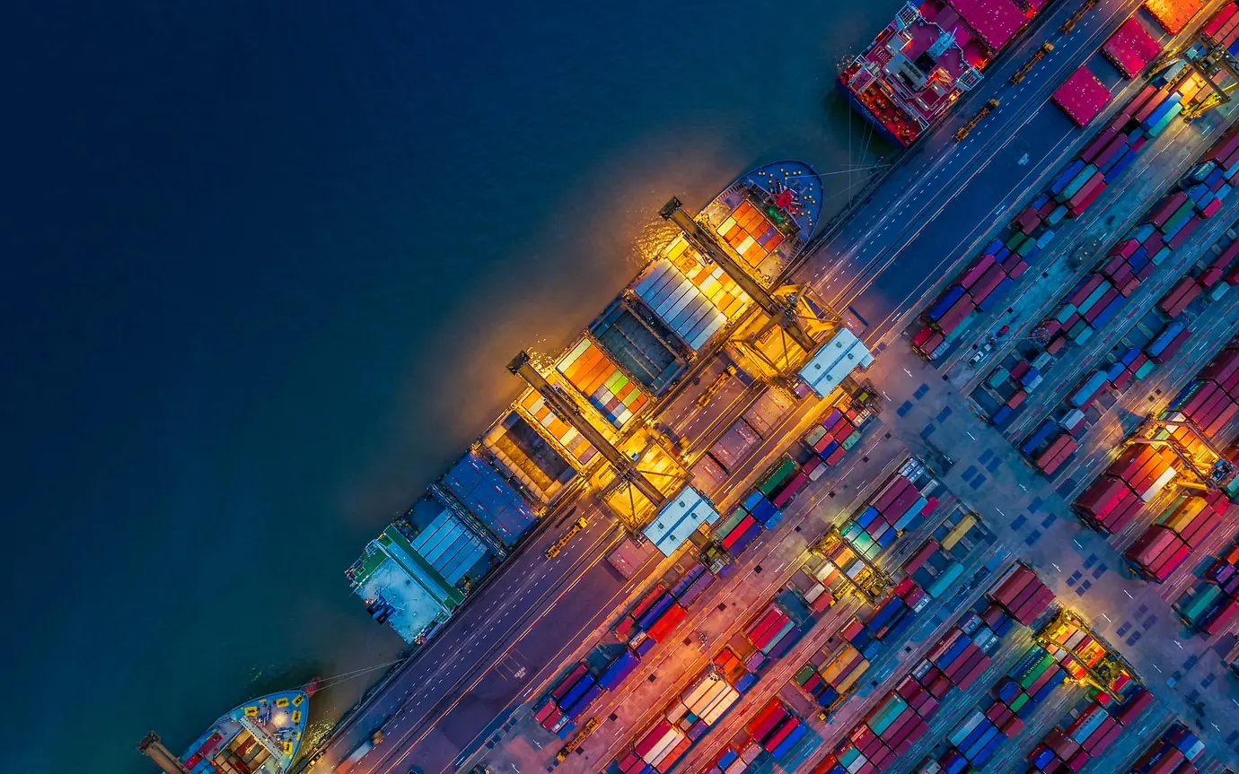 A dock covered in shipping containers in a variety of colours. A ship is docked and is being loaded at dusk.