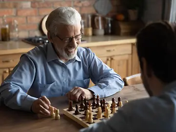 Smiling elderly Caucasian 60s father have fun play chess at home on weekend together with adult son.