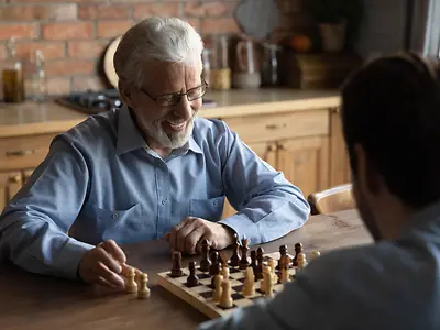 Smiling elderly Caucasian 60s father have fun play chess at home on weekend together with adult son.