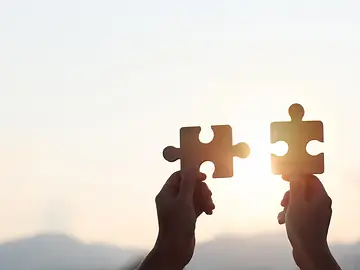 Hands are raised against a bright, white sky, each holding a piece of a wooden jigsaw puzzle.